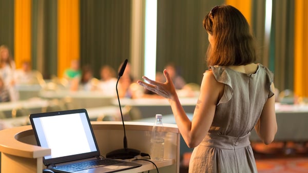 woman presenting to a large group in front of a microphone and her laptop.